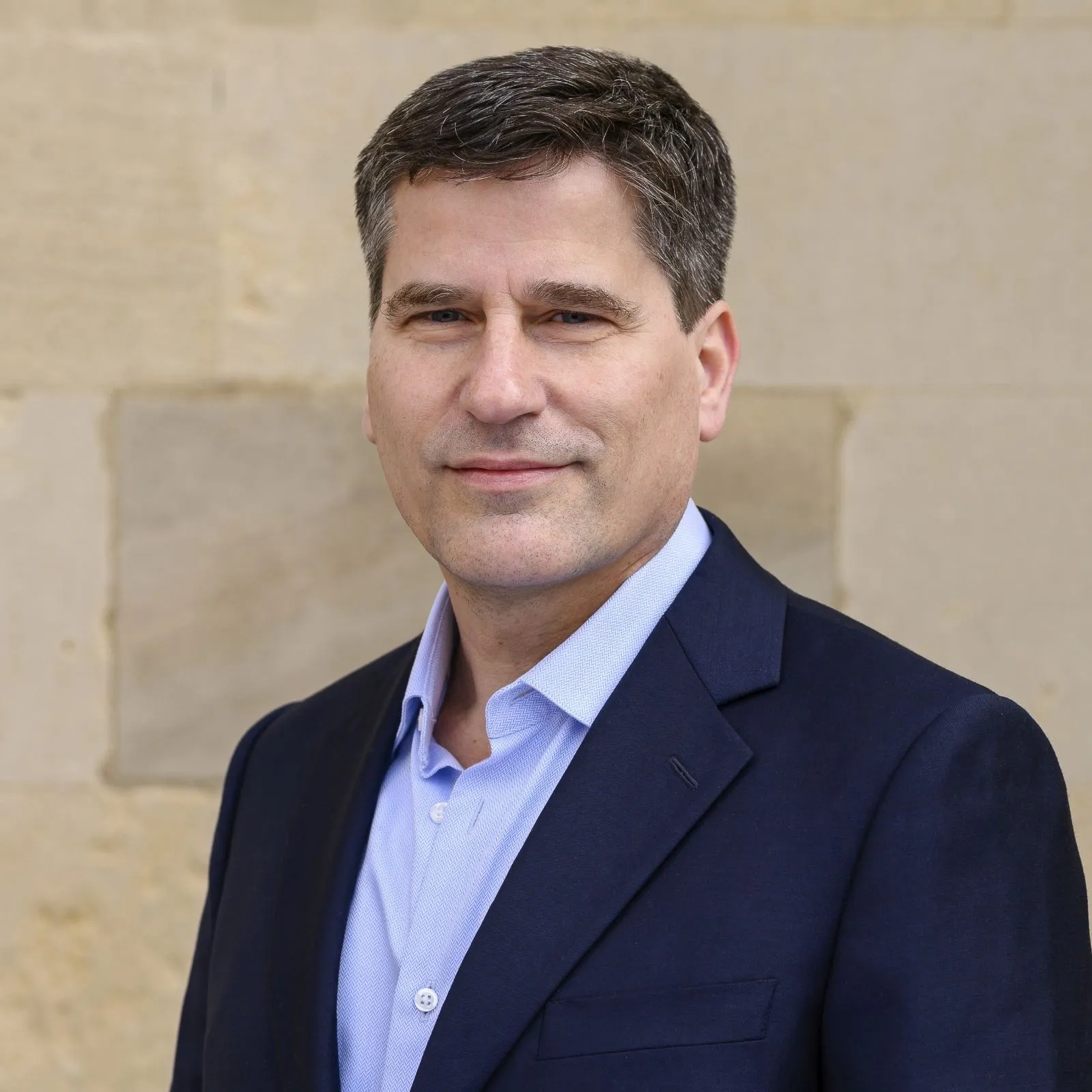 Portrait of a middle-aged man with short dark hair, wearing a navy blazer and light blue shirt, standing against a beige stone wall.