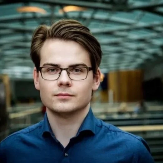 A young man with brown hair and glasses, wearing a blue button-up shirt, standing outdoors with a blurred background of a modern structure with glass and metal elements.