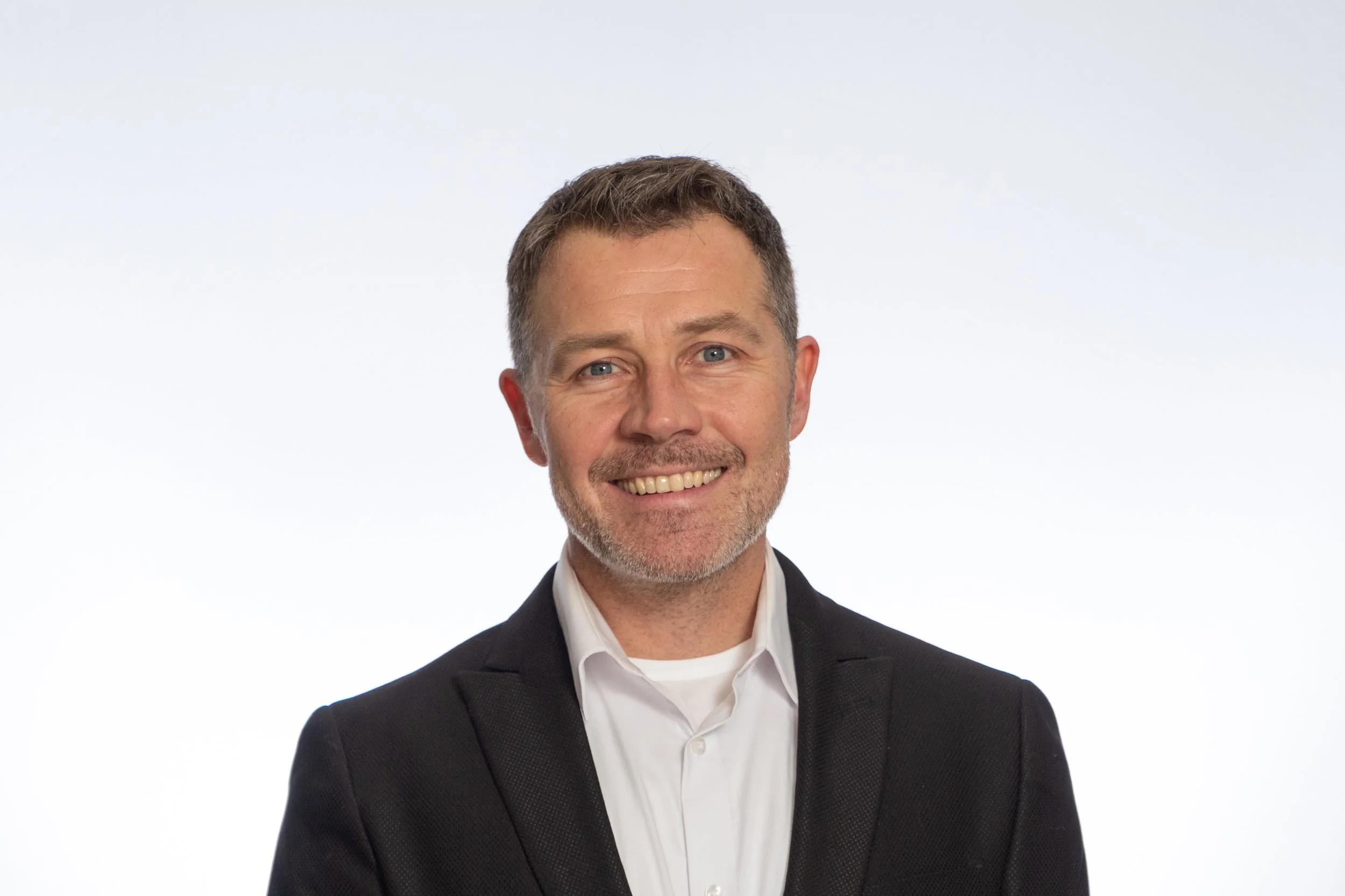Portrait of Daniel Omar Frimannson, Axelyf's Head of Nucleic Acids and Biology; a smiling middle-aged man with short dark hair and a beard, wearing a black suit jacket and white shirt, standing against a plain white background.