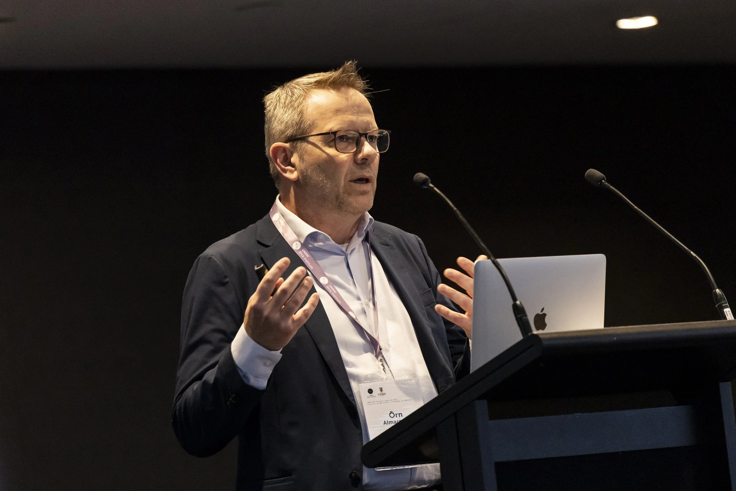 A man in a suit and glasses standing at a podium giving a speech, with a laptop and two microphones in front of him.