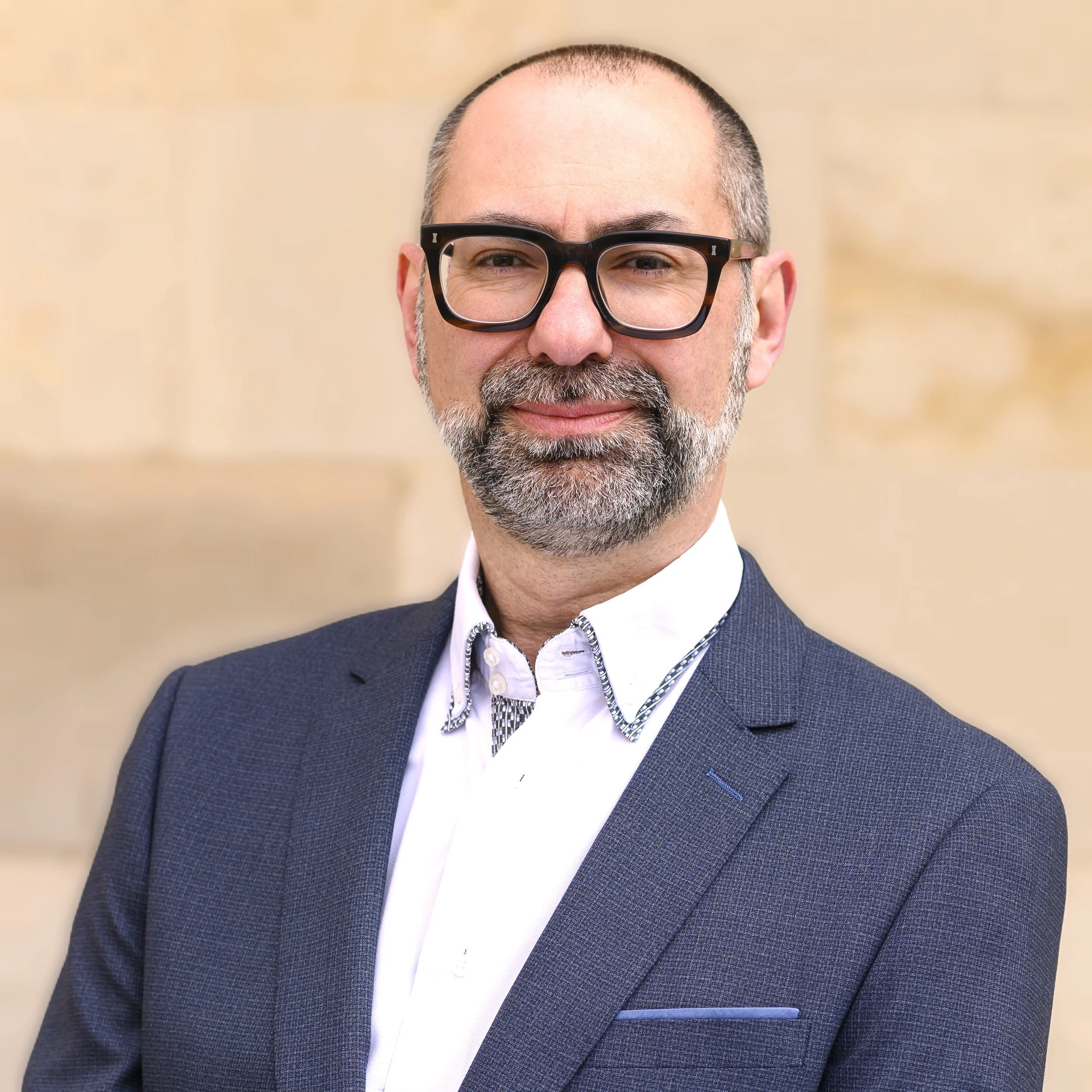 A middle-aged man with a beard and glasses, wearing a dark suit jacket and a white dress shirt, smiling at the camera.
