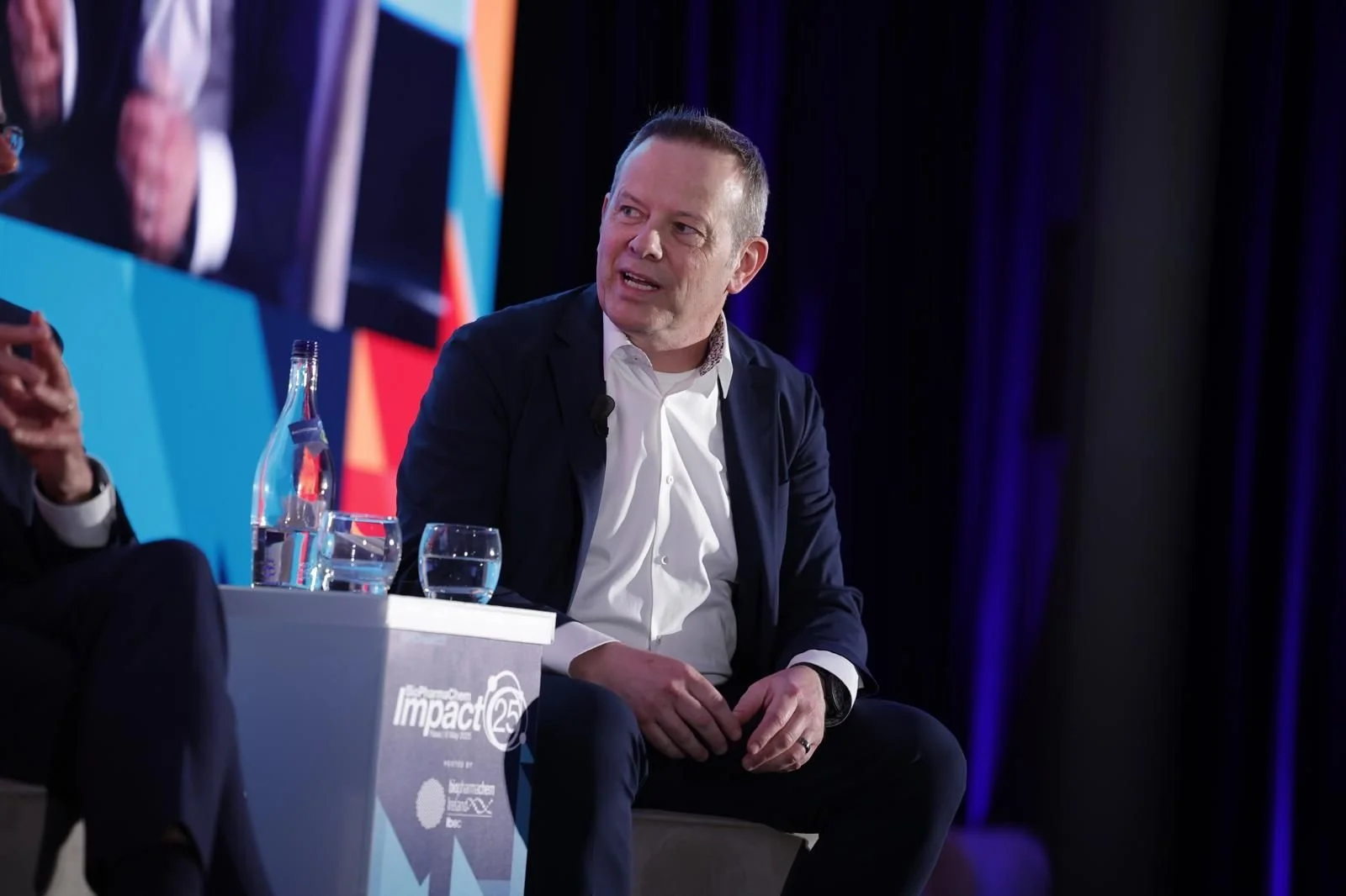 A man in a dark suit and white shirt sitting on a stage during a panel discussion. There are water bottles and glasses on a table in front of him. A large screen displays part of his image behind him, with colorful banners in the background.